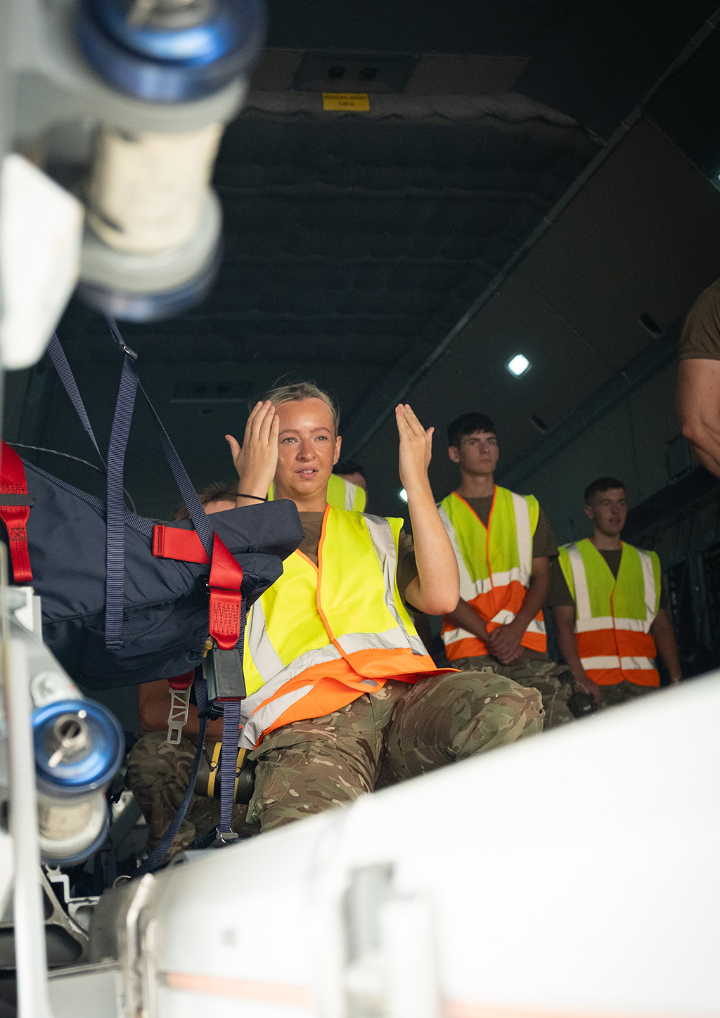 A member of 1 Air Mobility Wing, wearing a high-visibility vest, guides the driver of the Condec to the aircraft with her hands, while seated inside its dark cargo hold. Other 1 Air Mobility Wing personnel are visible behind her, ready to load the pallet on the Atlas.